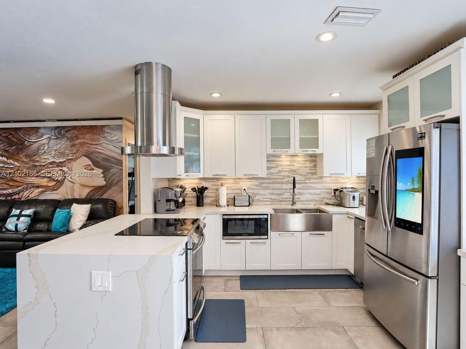 Kitchen featuring white cabinetry with glass-front uppers, waterfall-edge stone island, stainless steel cylindrical range hood, farm-style sink, and stone-tile flooring