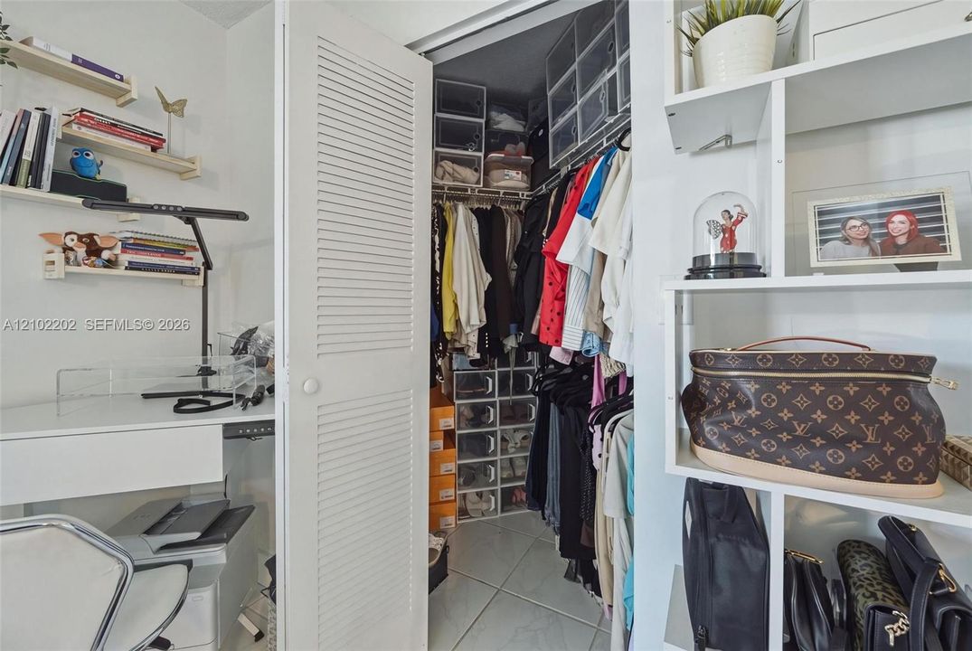 Integrated white shelving, louvered closet door, built-in closet organization, white desk surface, and neutral tile flooring