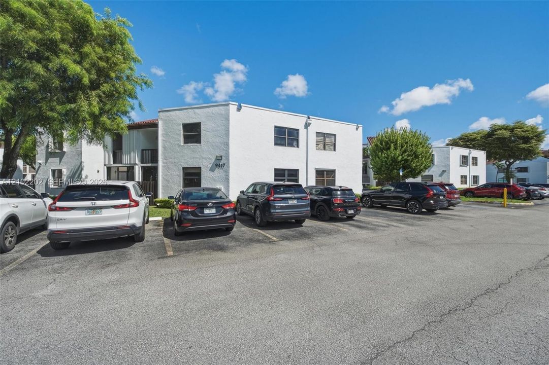White stucco exterior with multiple windows, surrounded by mature trees, and an asphalt parking area