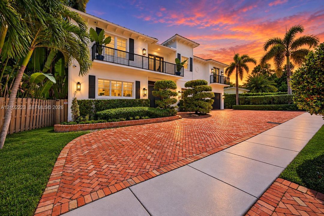 Two-story exterior featuring 2 car garage, circular drive, black balcony railings, a herringbone-patterned brick driveway, manicured landscaping, and outdoor wall mounted lighting