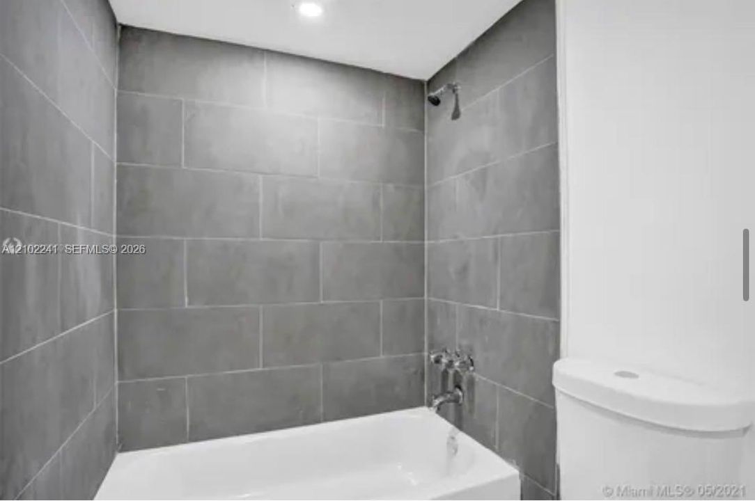 Full-height gray ceramic wall tiling, white porcelain bathtub, chrome wall-mounted shower head, chrome faucet fixtures, and white toilet