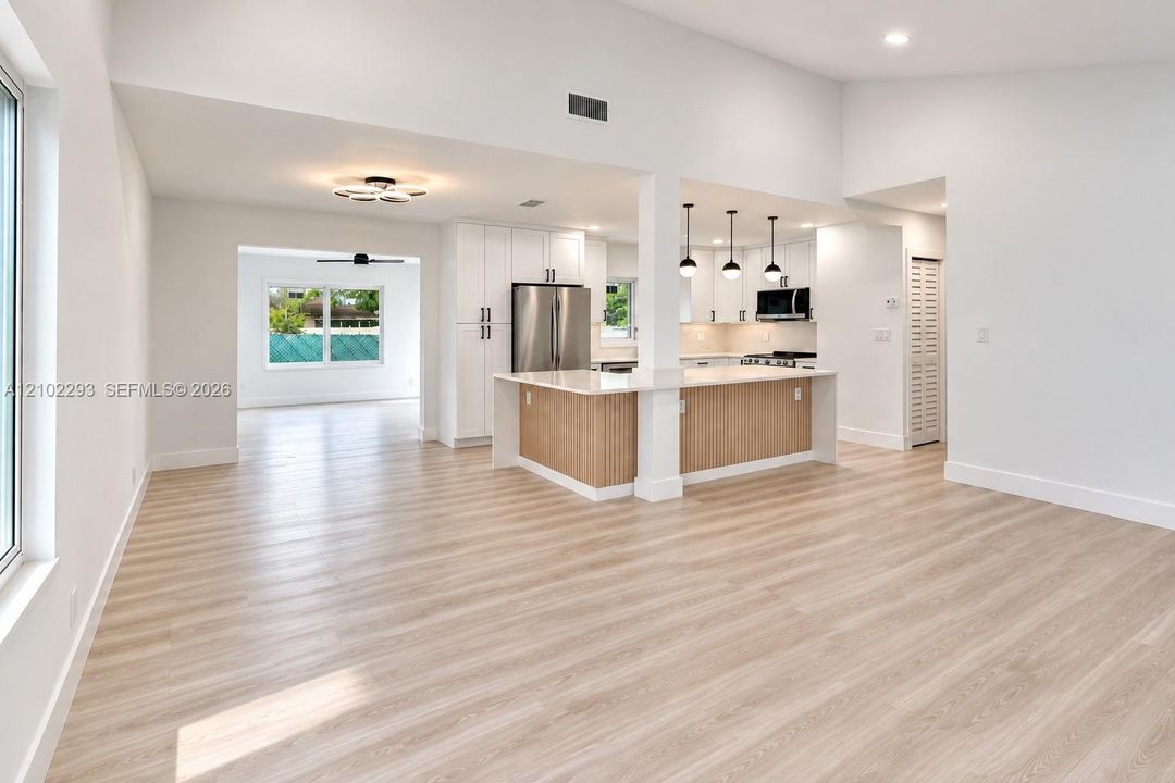 Open-concept interior featuring vaulted ceilings, light wood-finish flooring, a kitchen island with stone countertops, white shaker-style cabinetry, and pendant lighting