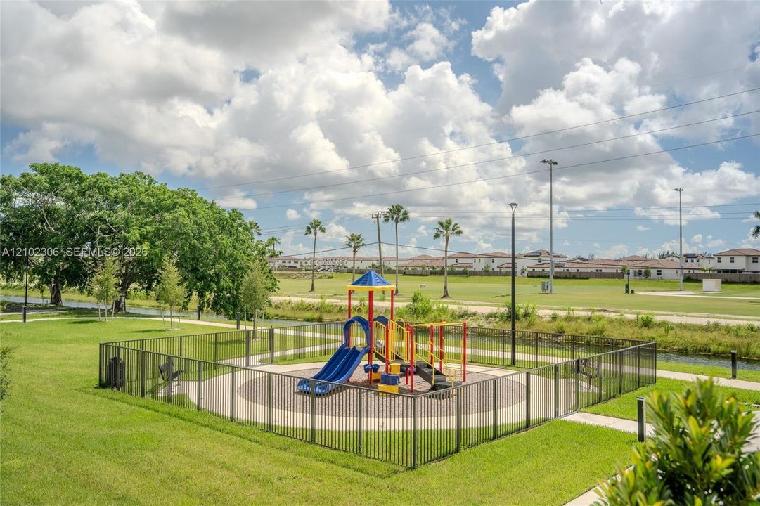 Outdoor recreation space featuring a fenced playground, manicured green space, paved pathways, and mature trees