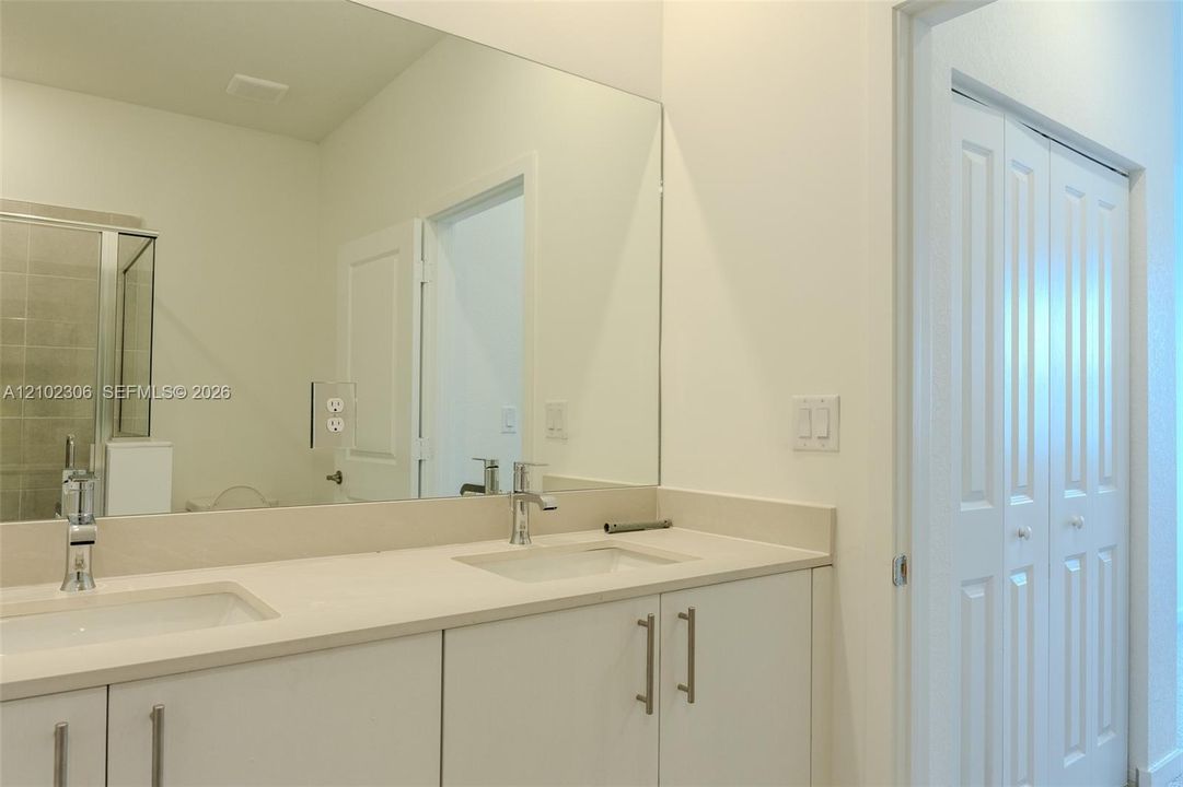 Dual-sink vanity with a solid-surface countertop, chrome faucets, floor-to-ceiling mirror, and white shaker-style cabinetry with bi-fold closet doors
