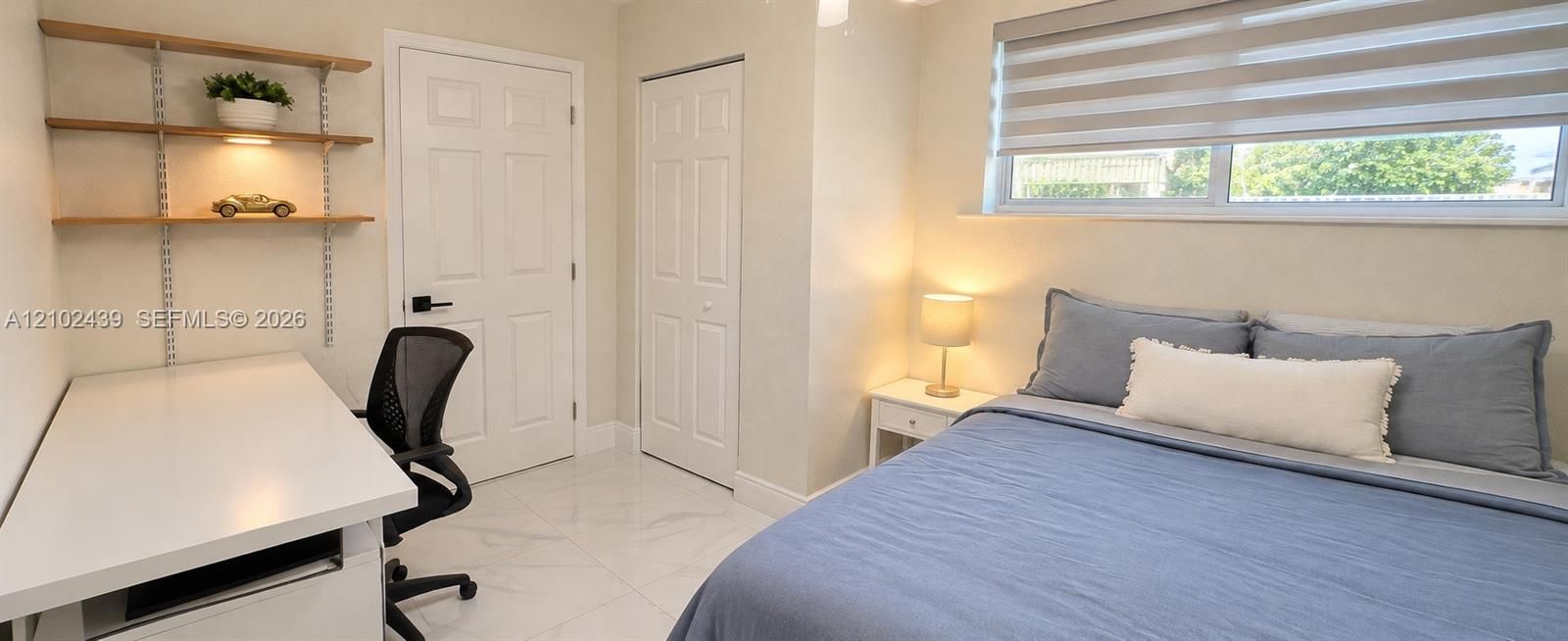 White tile flooring throughout, built-in shelving with integrated lighting, a solid panel interior door, a bi-fold closet door, and a horizontal window with blinds