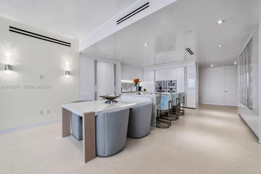 Modern kitchen featuring a central island with a stone countertop, integrated high-end stainless steel appliances, seamless white cabinetry, recessed ceiling lighting, and linear wall sconces