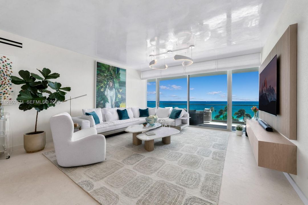 Living area featuring floor-to-ceiling glass sliding doors, high-gloss ceiling finish, custom wall-mounted wood-finish media console, and direct ocean backdrop