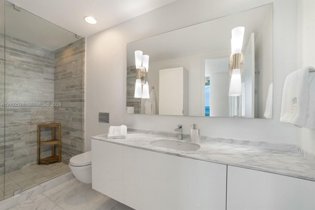 Floating vanity with integrated sink, stone-surface countertop, walk-in glass-enclosed shower featuring horizontal stone-tiled walls, wall-mounted toilet, and dual decorative light fixtures