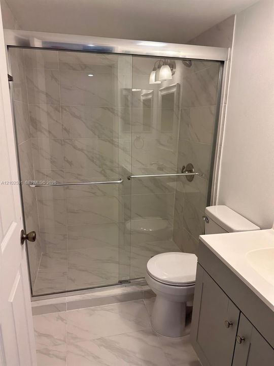 Shower featuring a frameless glass sliding door, floor-to-ceiling stone-look tiling, vanity with solid-surface countertop, and satin nickel hardware