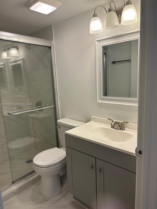 Grey vanity cabinet with a white countertop and integrated sink, framed vanity mirror, three-light fixture, glass-enclosed shower with tiled surround, and tiled flooring