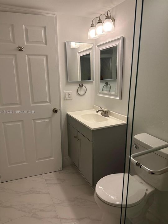 Bathroom featuring large-format tile flooring, a gray vanity cabinet with an integrated white sink, a framed wall mirror, a three-light vanity fixture, and a glass-enclosed shower
