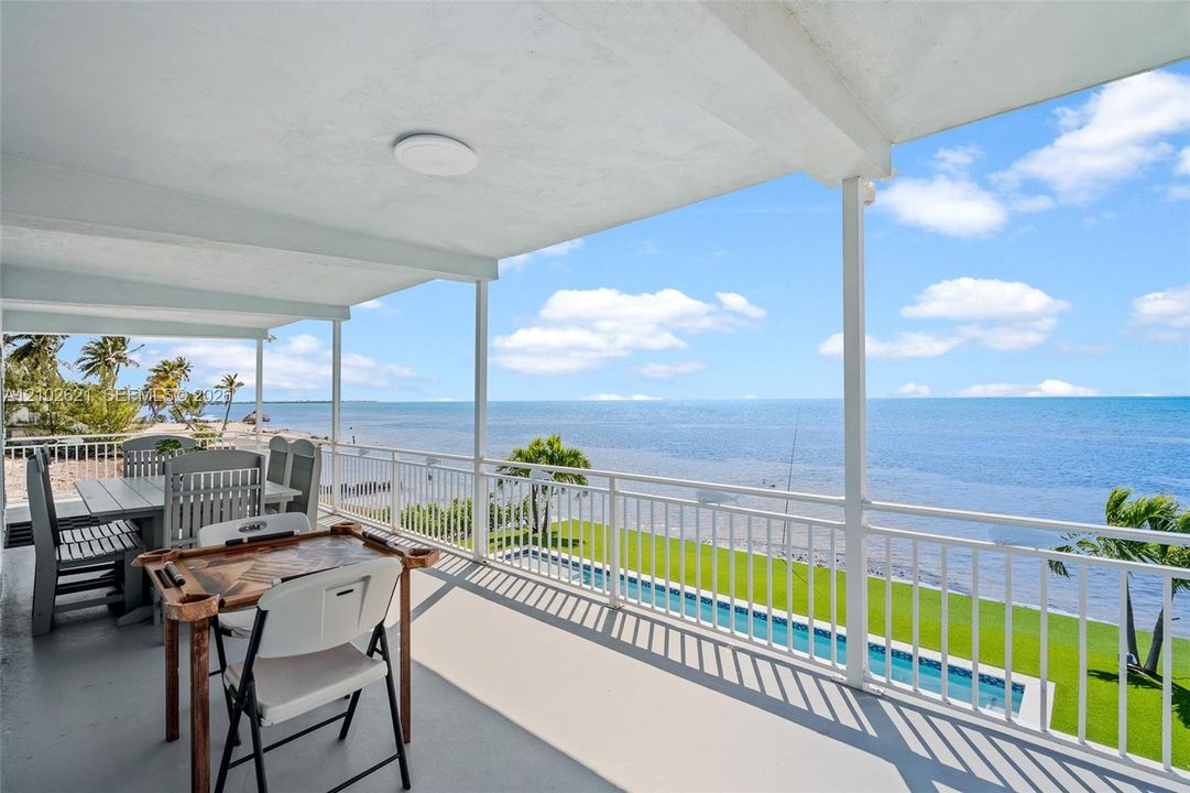 Covered exterior balcony with a white railing, overlooking an in-ground swimming pool and expansive body of water