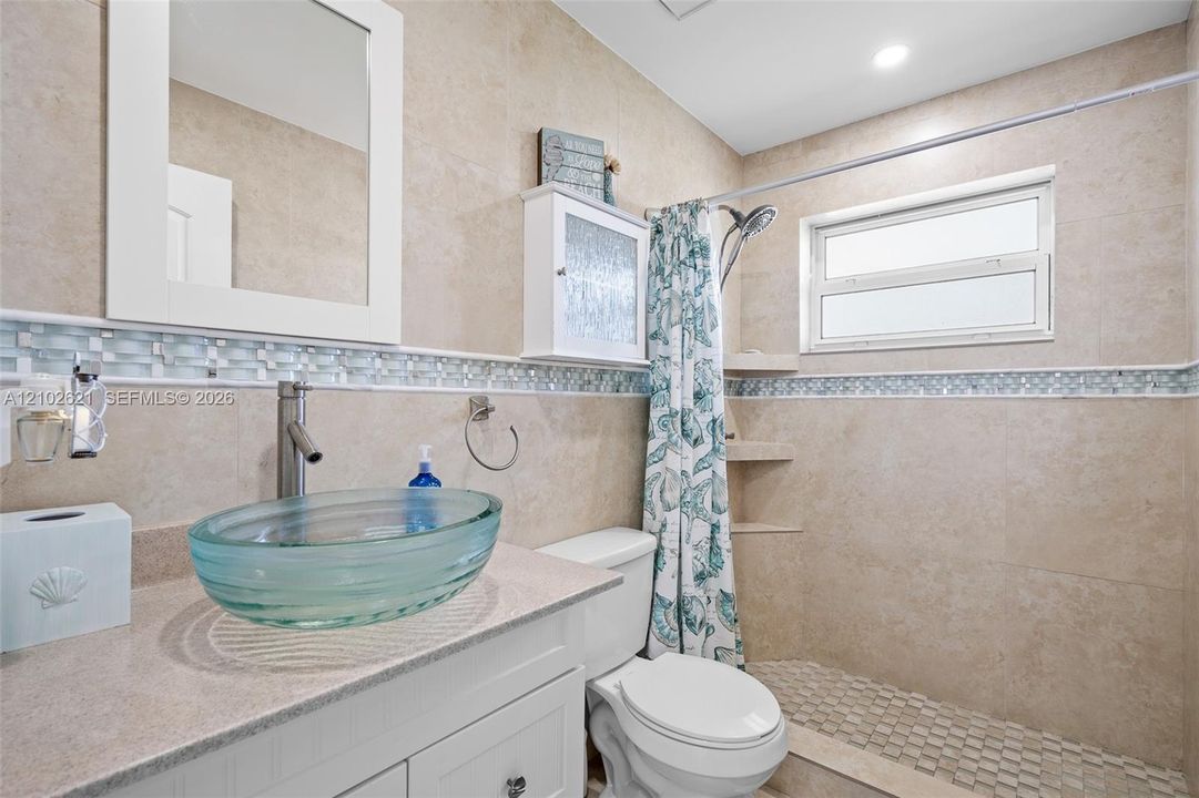 Glass vessel sink, light-toned countertop, vanity with cabinet storage, tiled shower with recessed shelves, and an operable window