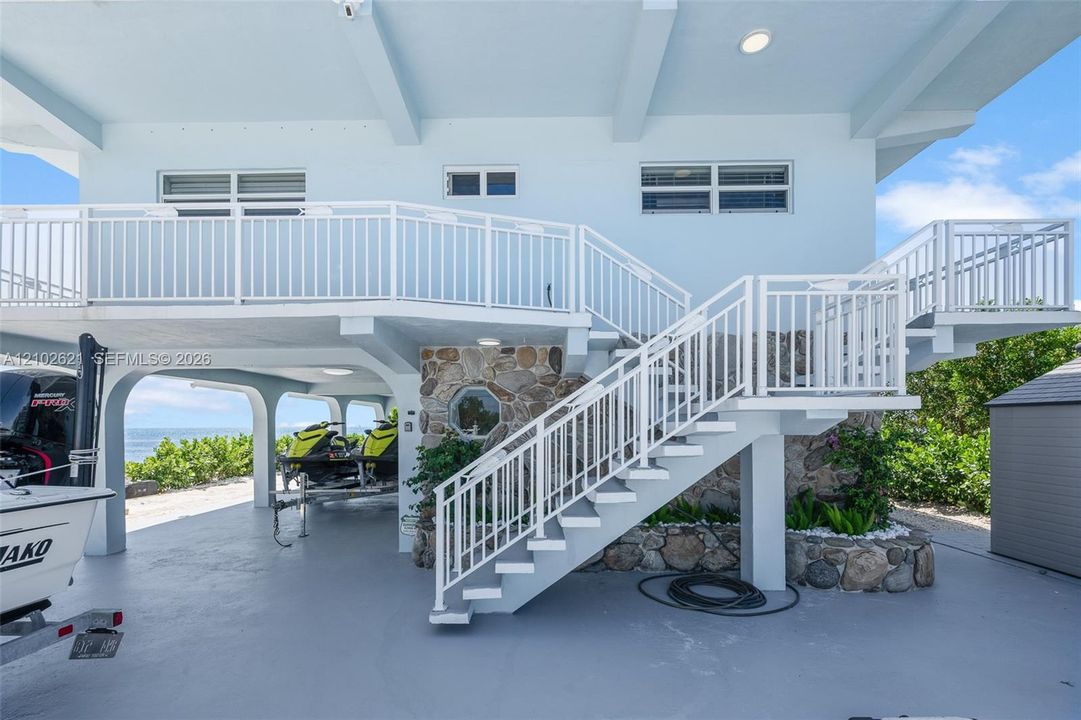 Covered parking area with concrete flooring, dual exterior staircases with white railings, and stone-clad accents