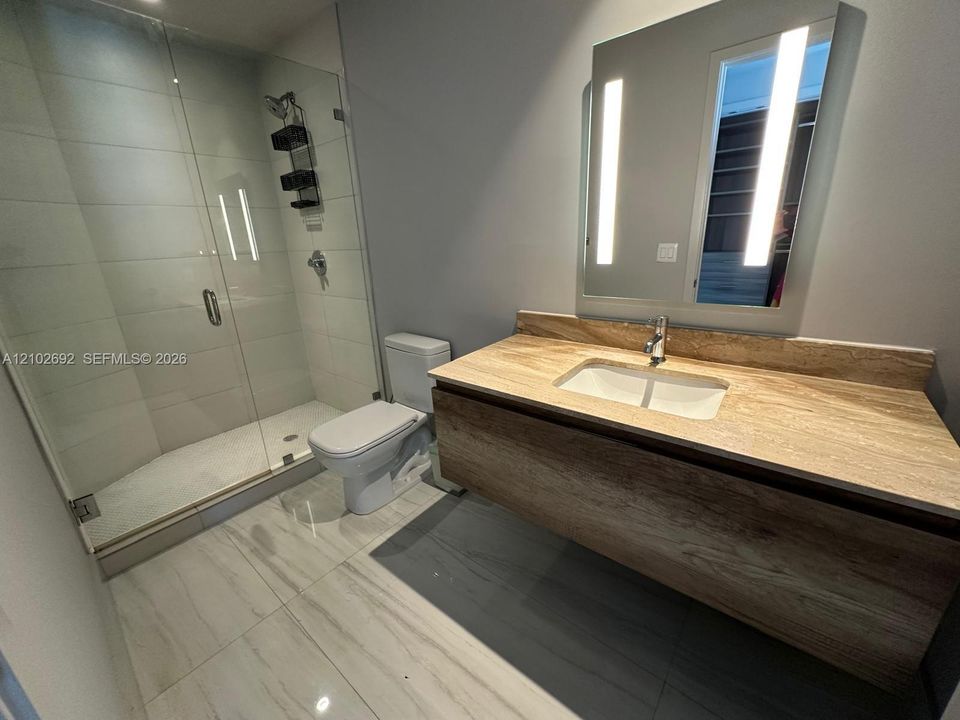 Floating wood-finish vanity with an integrated countertop sink, wall-mounted mirror with integrated lighting, glass-enclosed shower, and large-format tile flooring