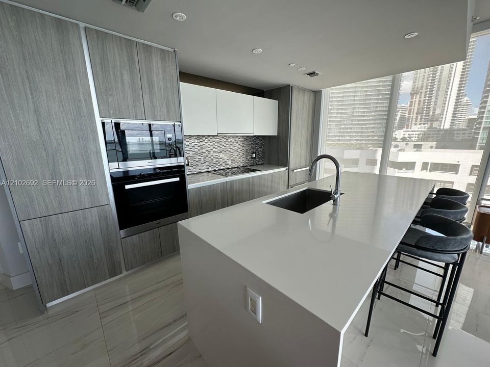 Kitchen featuring a central quartz-top island with an integrated sink, custom wood-finish cabinetry, built-in stainless steel appliances, and floor-to-ceiling windows