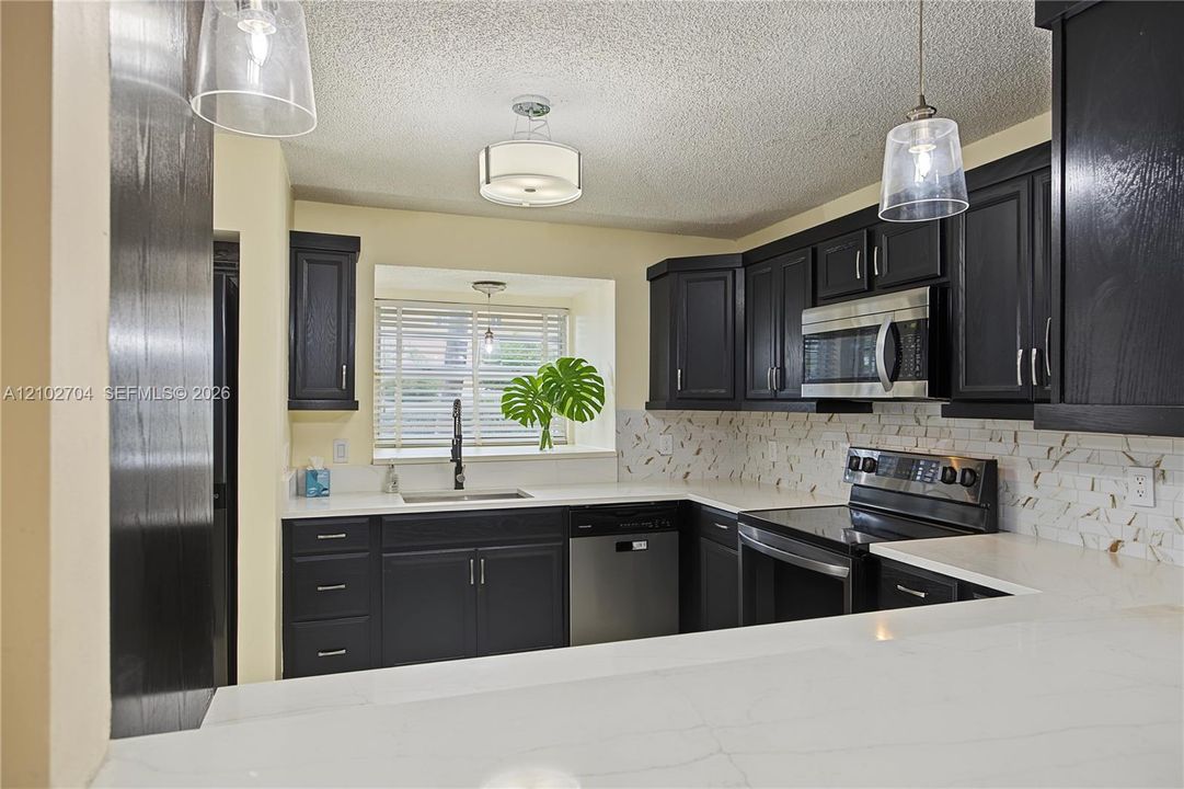 Kitchen featuring dark shaker cabinetry, light-toned stone-finish countertops, stainless steel appliances, patterned tile backsplash, and two glass pendant lights