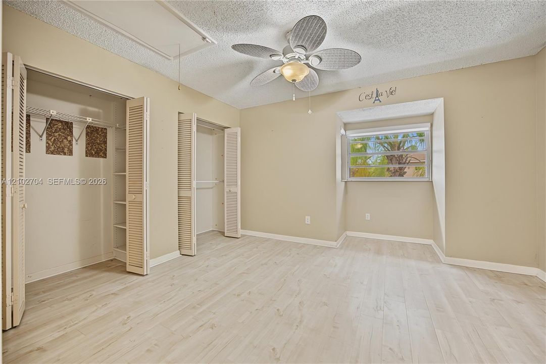 Light-toned wood-finish flooring, two closets with louvered doors and built-in shelving, a ceiling fan with integrated lighting, and a recessed window nook