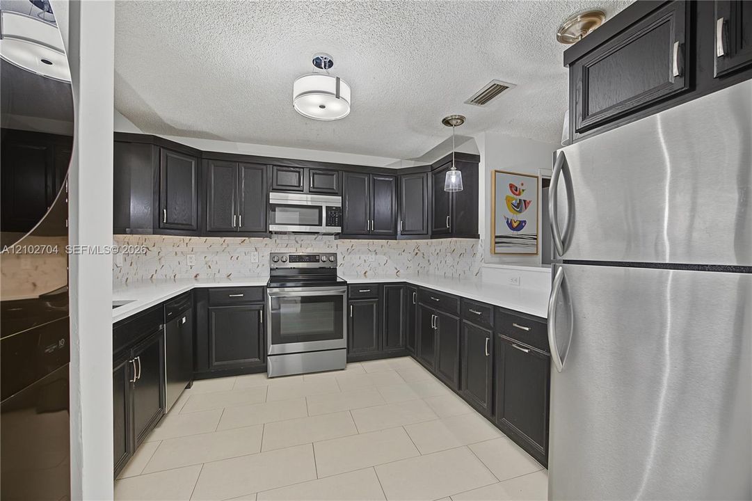 Kitchen featuring dark-finish cabinetry, white countertops, a patterned tile backsplash, stainless steel appliances, and large-format tile flooring