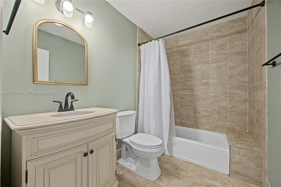 Bathroom featuring a vanity with a stone countertop and under-mount sink, matte black faucet hardware, arched wall mirror, light fixture with three glass shades, and a tile-surround shower and tub combination