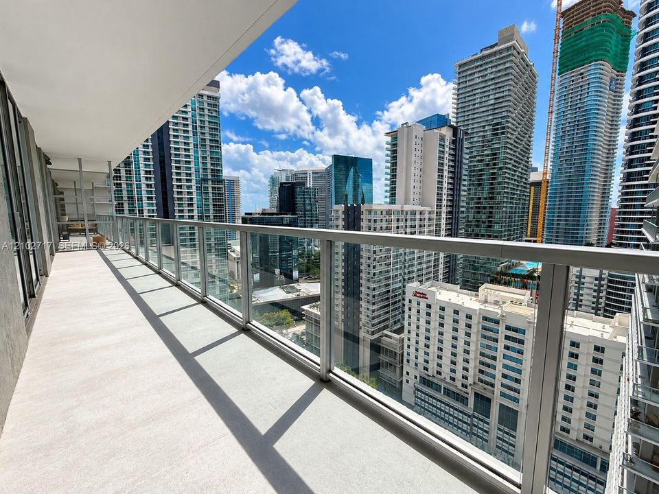 Expansive balcony featuring concrete flooring, a glass panel railing with metallic frame, and high-rise urban scenery
