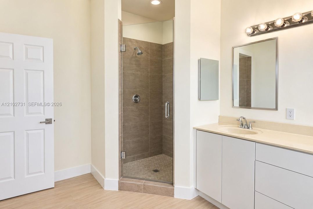 Enclosed shower with full-height tile surround and glass door, integrated vanity with solid surface countertop, rectangular wall-mounted mirror, and multi-bulb polished finish vanity lighting
