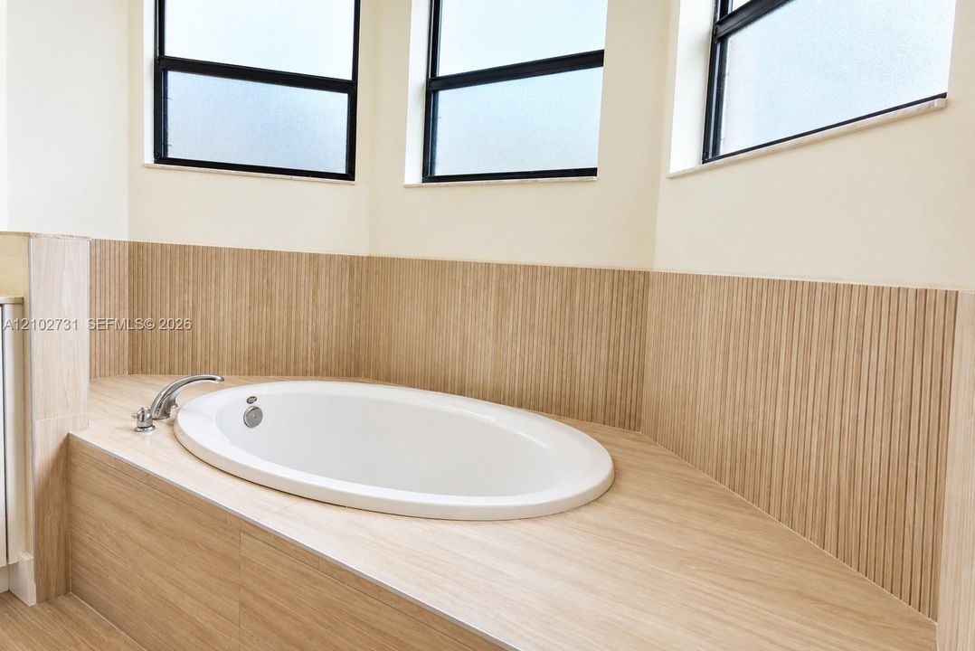 Oval soaking tub featuring vertical textured tile surround, horizontal tile decking, and metal fixtures set beneath triple windows with black frames