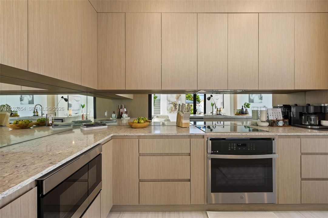 Modern kitchen featuring flat-panel wood-finish cabinetry, neutral stone countertops, a mirrored backsplash, built-in stainless steel oven, and a flat-surface cooktop