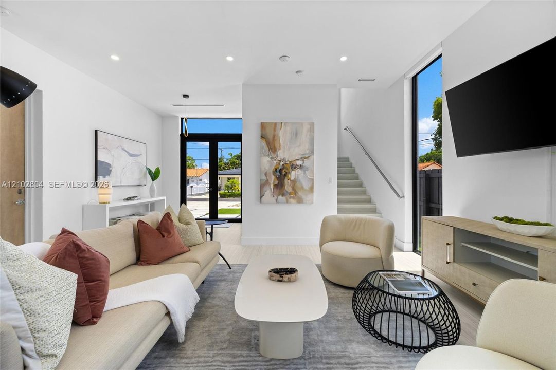 Open-concept living space featuring high ceilings, recessed lighting, full-height windows, a glass-paned entry door, and a floating staircase with a metal handrail