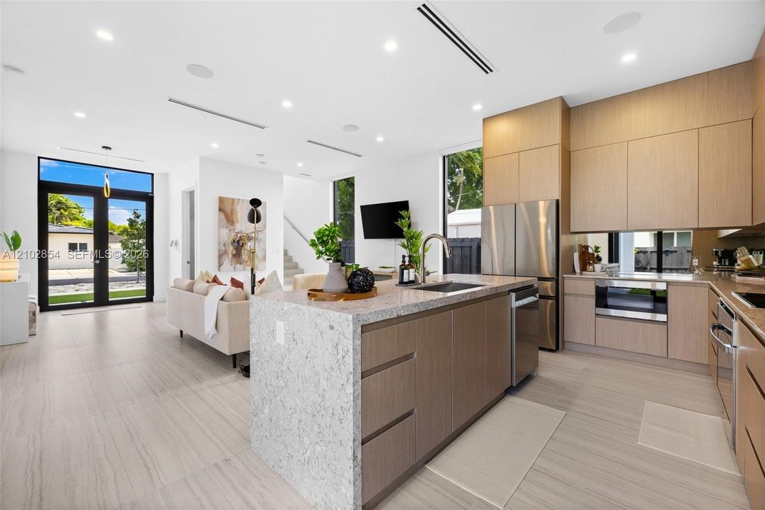 Modern kitchen featuring flat-panel wood cabinetry, a waterfall-edge center island with stone countertops, stainless steel appliances, and large-format tile flooring