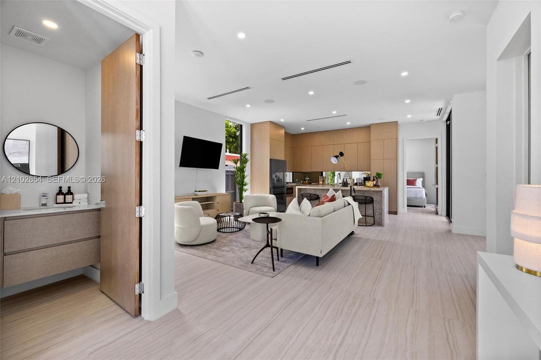 Open-concept interior featuring light wood-finish flooring, recessed lighting, kitchen cabinetry with flat-panel wood-finish doors, a center island with stone-finish waterfall countertop, and a vanity with a circular mirror