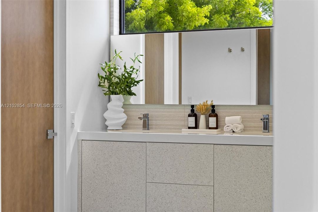Floating vanity with textured cabinetry, solid surface countertop, linear tile backsplash, horizontal mirror, and chrome dual-faucet fixtures
