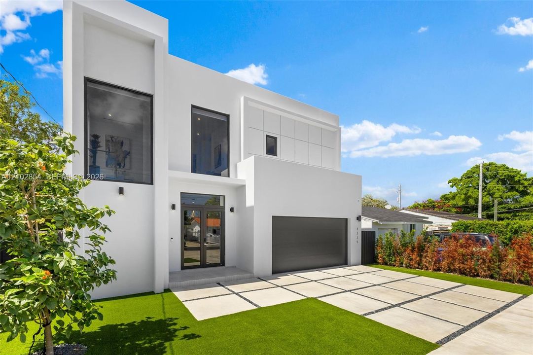 Modern architectural exterior featuring a smooth white stucco facade, expansive floor-to-ceiling windows, a dark metal garage door, and a geometric concrete paver driveway with artificial turf inlays