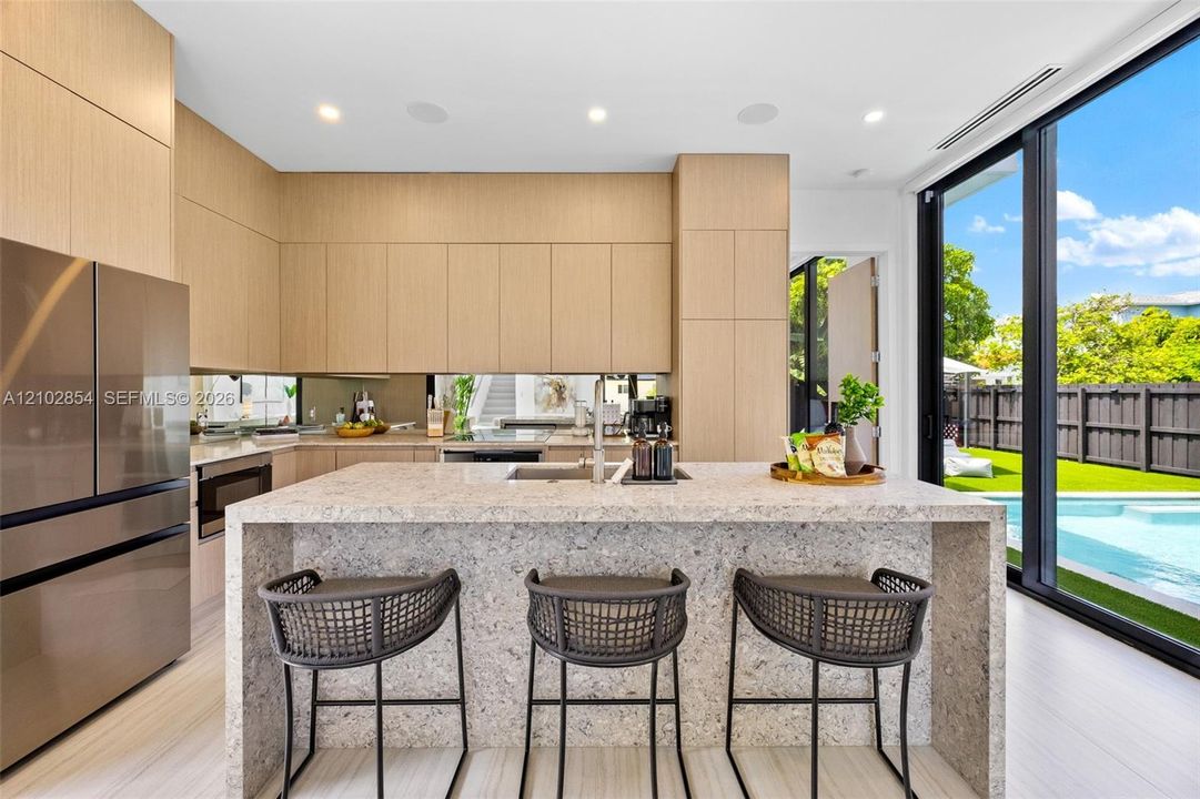 Flat-panel cabinetry, waterfall-edge stone island with integrated sink, floor-to-ceiling glass sliding doors, stainless steel refrigerator, and recessed lighting
