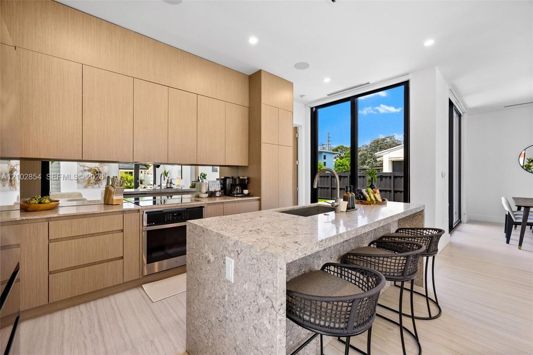Kitchen featuring flat-panel wood-finish cabinetry, stone waterfall-edge island, mirrored backsplash, built-in wall oven, and floor-to-ceiling windows