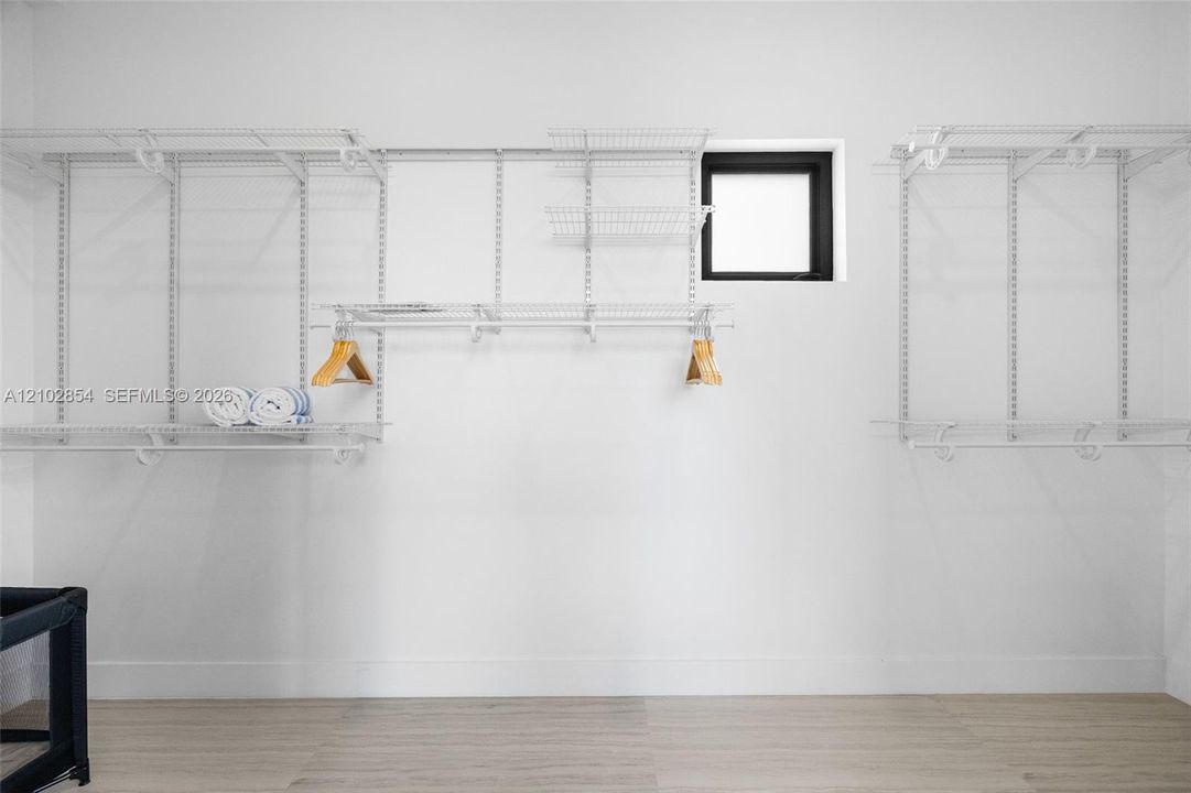 Customizable white wire shelving system featuring multiple hanging rods and tiered organizers, wood-finish flooring, and a framed window with a black finish