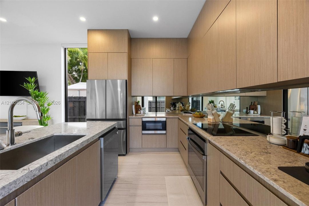 Contemporary kitchen featuring full-height flat-panel cabinetry, light stone-finish countertops, stainless steel appliances, mirror backsplash, and recessed lighting