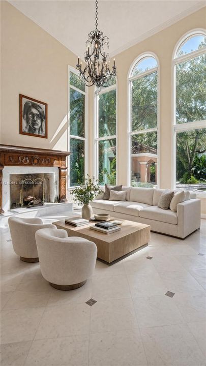 High ceilings featuring an ornate chandelier, a fireplace with a carved wood surround, arched windows, and light-toned tile flooring with dark accent inlays