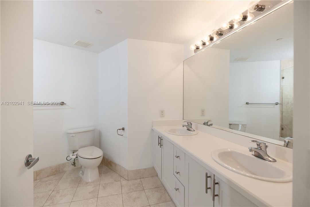 Bathroom featuring a dual-sink vanity with white cabinetry, light-toned countertops, a full-width wall mirror, and neutral-toned tile flooring