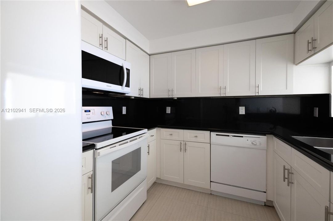 Kitchen featuring white shaker-style cabinetry, black stone countertops, matching black backsplash, and white appliances including a built-in microwave and dishwasher