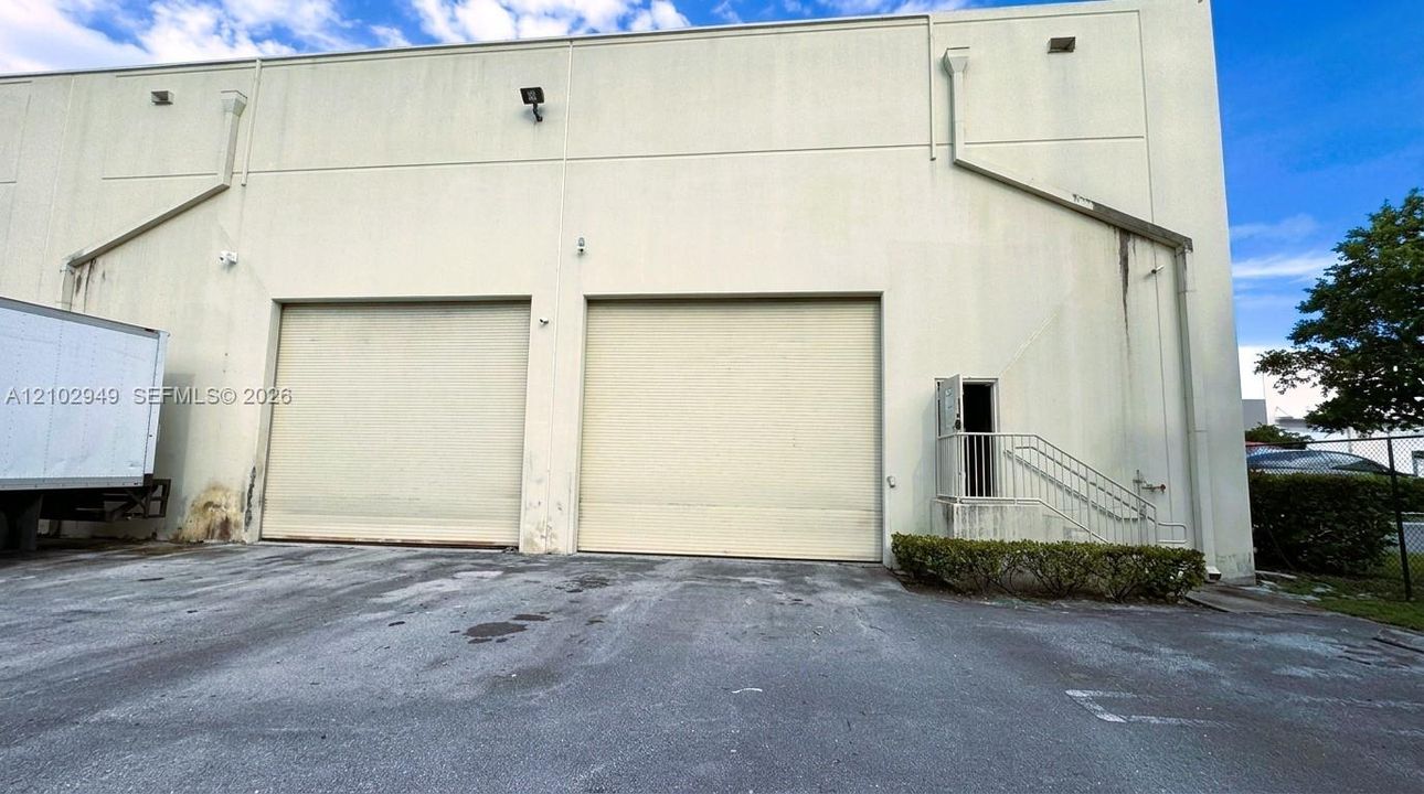 Industrial exterior featuring two high-clearance roll-up bay doors, concrete paved lot, and a metal staircase with integrated service entry