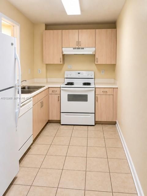 Kitchen featuring light wood-finish cabinetry, tile flooring, white appliances, and a double-basin sink with solid-surface countertops