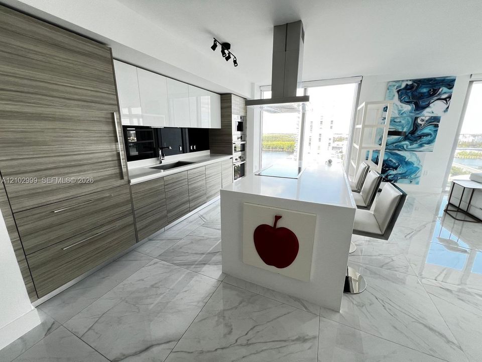 Contemporary kitchen featuring stainless steel appliances, white cabinetry with horizontal wood-finish accents, a central island with waterfall countertop, polished stone flooring, and a statement range hood