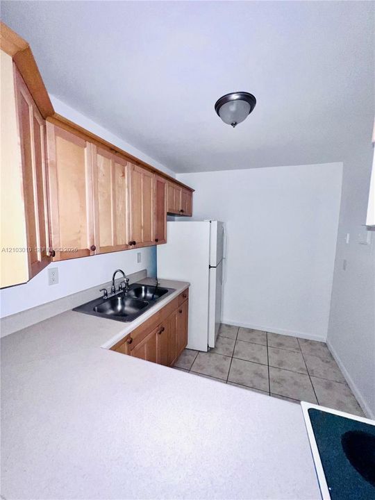 Kitchen featuring natural wood cabinetry, solid-surface countertops, dual-basin stainless steel sink, tile flooring, and integrated refrigerator