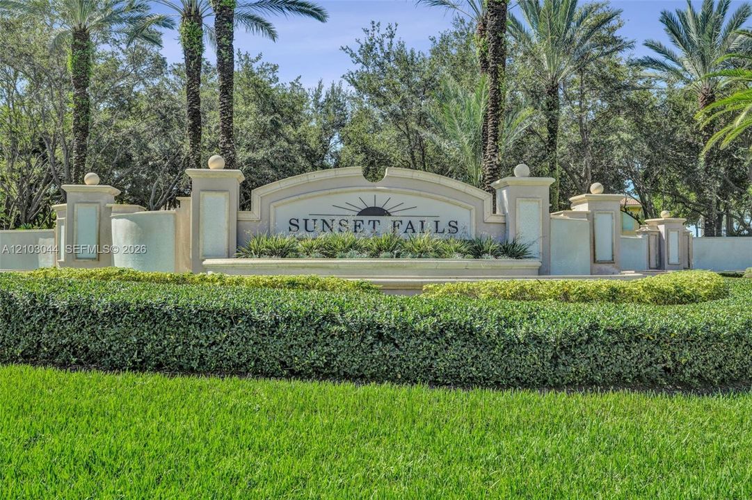 Stucco community signage featuring decorative masonry pillars, integrated lighting fixtures, tiered architectural detailing, and mature palm landscaping