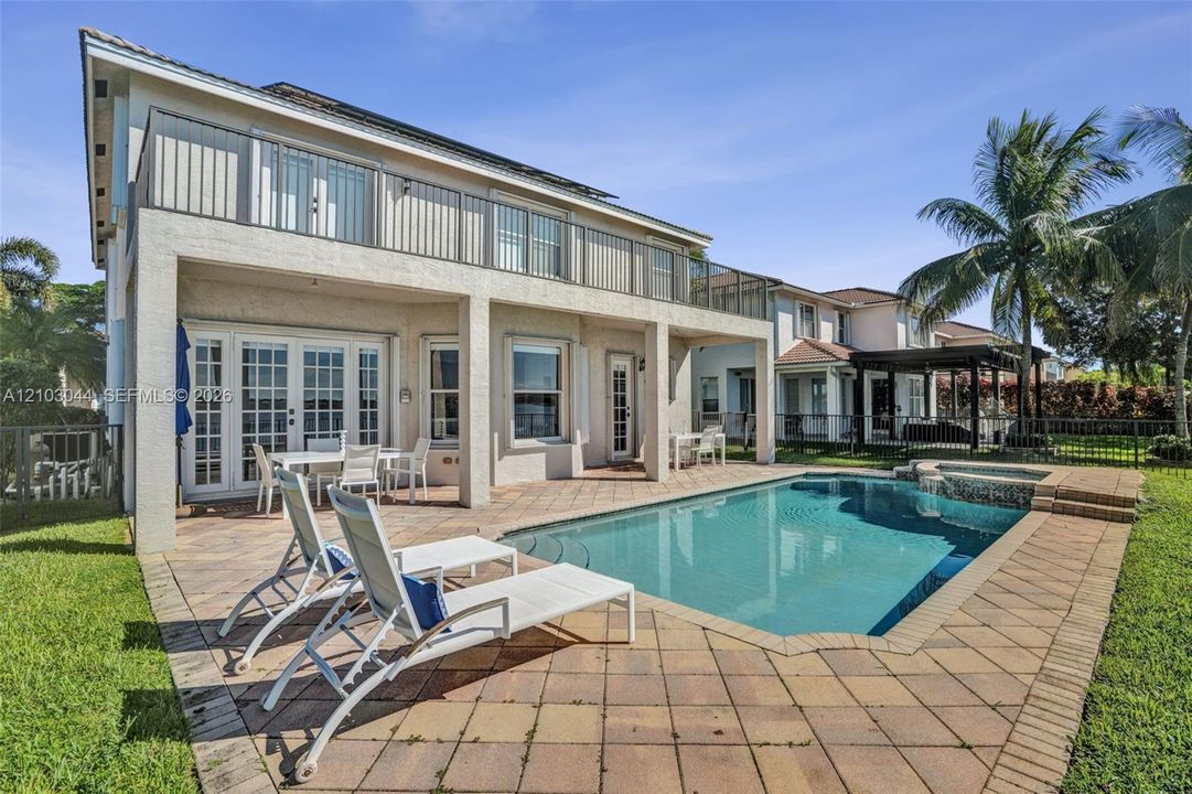 Stucco exterior featuring a second-story balcony with metal railings, a covered ground-level patio, and a concrete-paver pool deck surrounding an inground pool with an attached spa