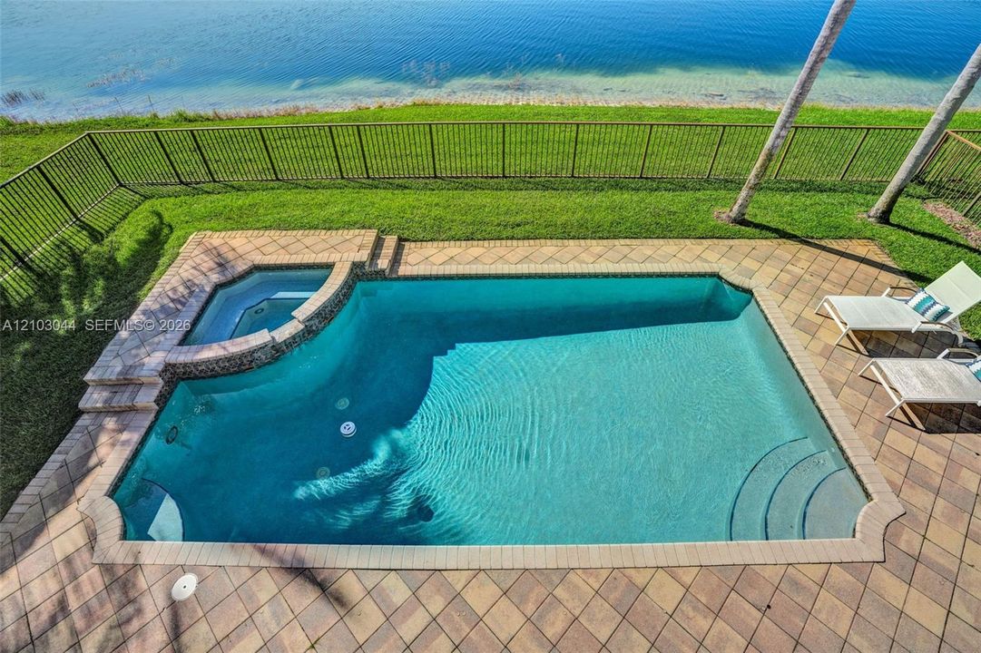 In-ground pool with an integrated spa, paver deck, metal fencing, and grass lawn bordering the water