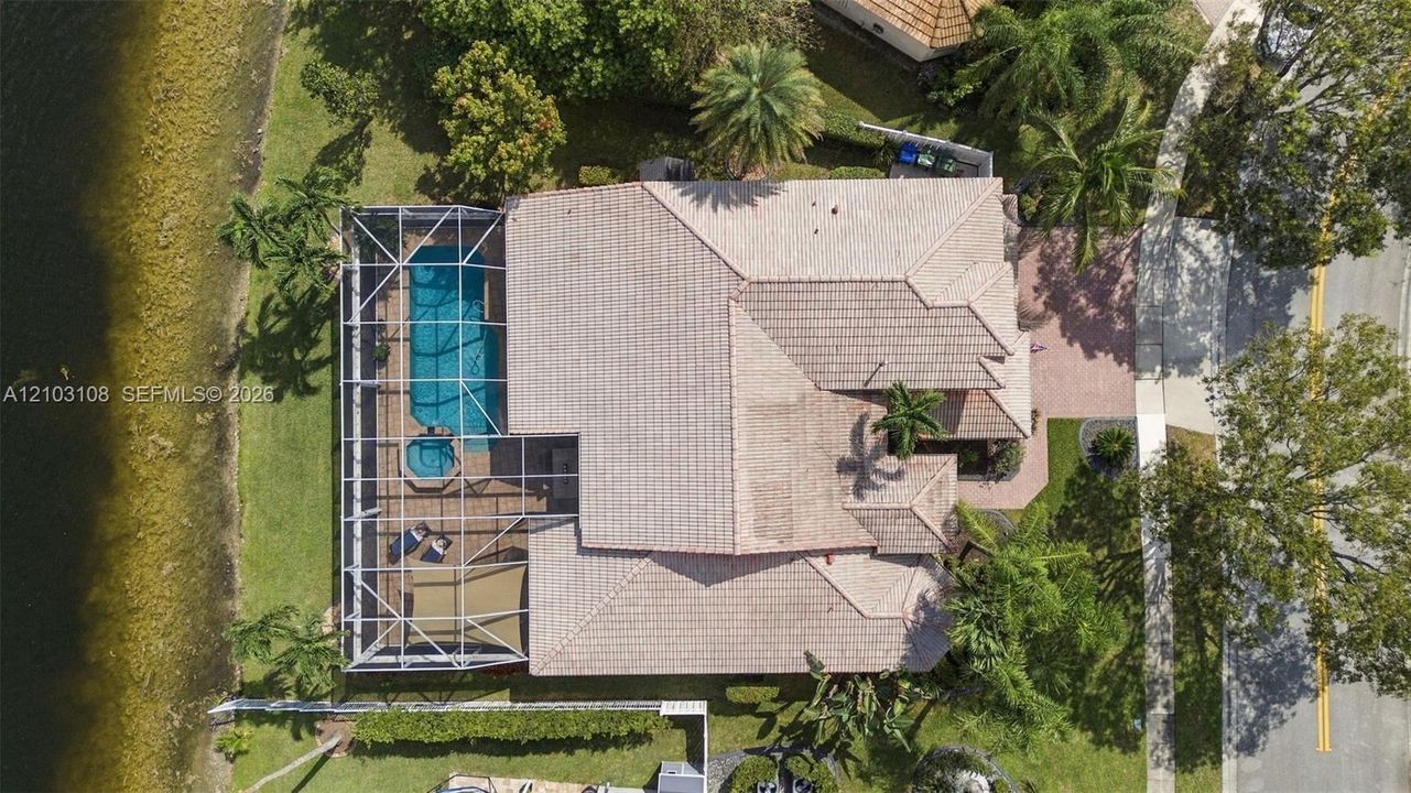 Aerial perspective featuring a tile roof, screen-enclosed pool and spa, paved driveway, and direct water frontage