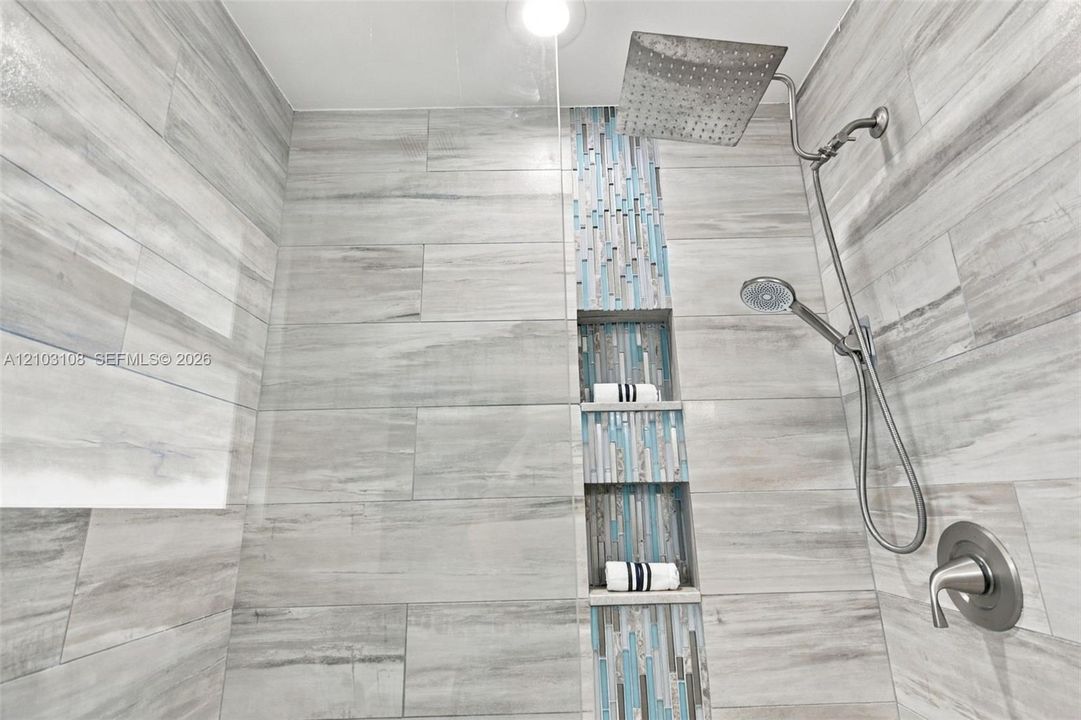 Walk-in shower featuring large-format ceramic tile, a vertical mosaic accent inlay, built-in tiered shelving, a rain-style showerhead, and a separate handheld sprayer