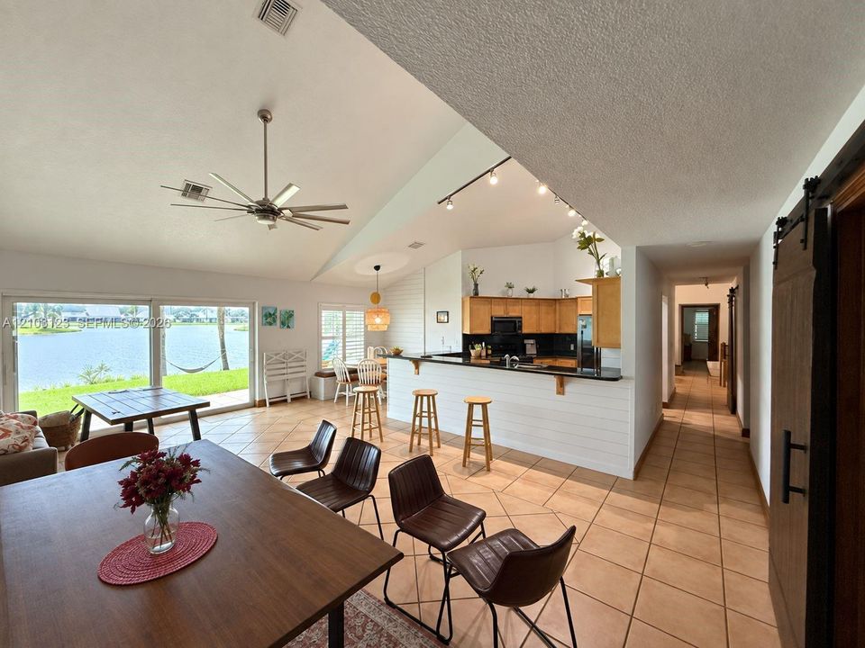 Open-concept living space featuring vaulted ceilings, a kitchen breakfast bar with white horizontal paneling, tile flooring, track lighting, and sliding glass doors with water views